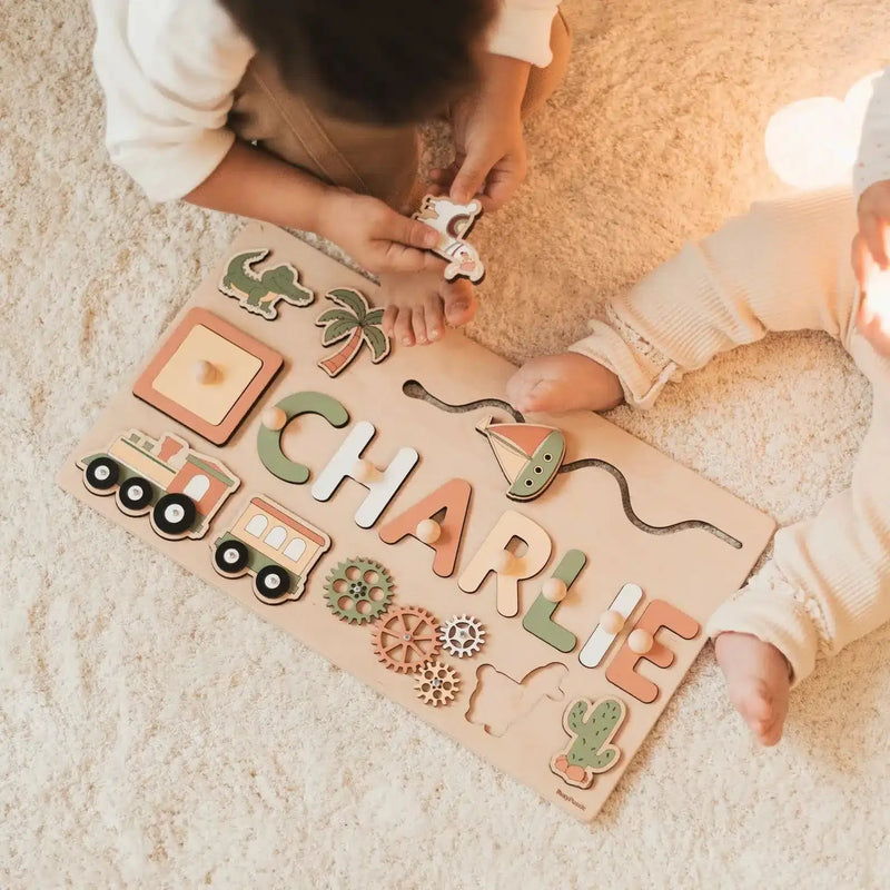 Deux enfants sont assis sur un tapis léger, jouant avec un Puzzle Personnalisé Prénom en Bois qui épelle « CHARLIE » et présente des formes amusantes comme un train, un cactus et un palmier.