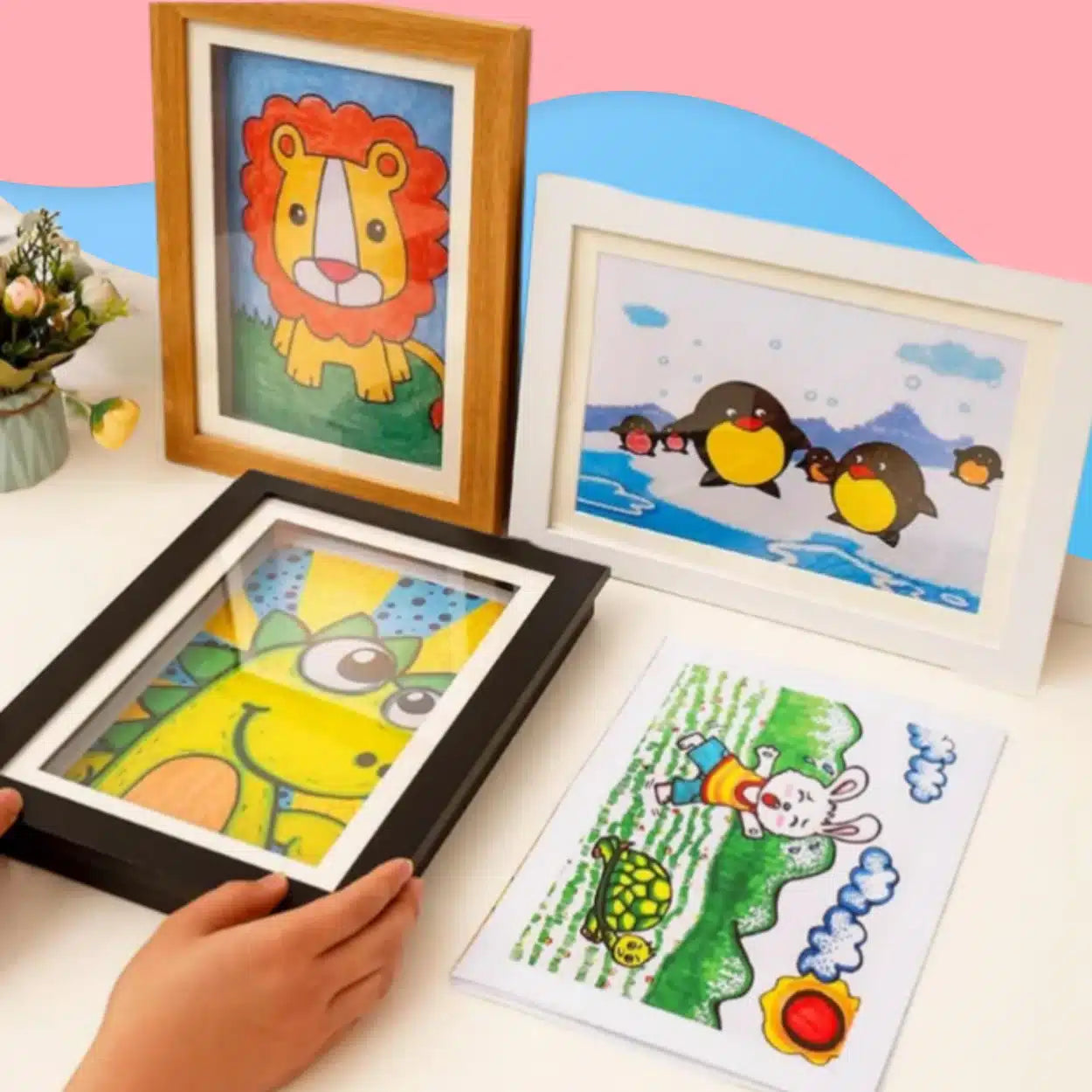 Quatre dessins d'animaux colorés en taille A4 (lion, pingouins, girafe, lapin avec tortue) exposés sur une table blanche avec un fond rose et bleu-parfait avec le Cadre pour dessins d'enfant pour stocker et exposer 150 œuvres.