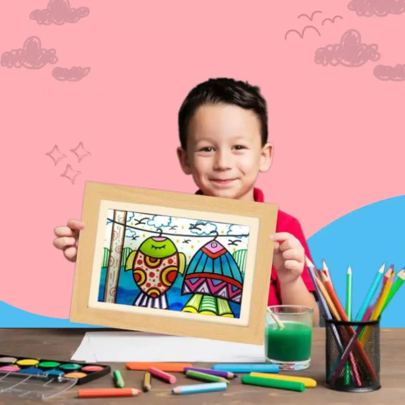 Un jeune garçon vêtu d'une chemise rouge est assis à une table avec du matériel d'artiste, montrant fièrement son dessin d'oiseau coloré dans le Cadre pour dessins d'enfant (A4), qui stocke et expose jusqu'à 150 œuvres d'art d'enfants. Fond rose et bleu.