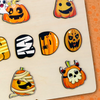Feuille d'autocollants décorée de visages de citrouilles de dessins animés sur le thème d'Halloween, y compris des momies, des fantômes et des crânes, disposés sur une surface en bois rustique - un souvenir parfait particulièrement adapté à la saison.