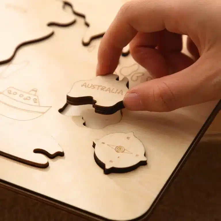 Une main place délicatement une pièce de puzzle en bois étiquetée « AUSTRALIE » dans le plateau Puzzle du monde en bois Montessori, avec une autre pièce à proximité.