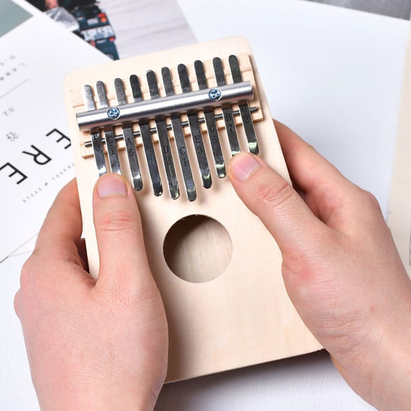 Mains jouant sur un kalimba en bois avec des dents en métal, positionnées sur une surface blanche.