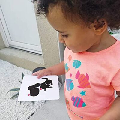 Un enfant portant une chemise rose ornée d'imprimés de poissons colorés tient un « Jouet montessori imagier carte pour les bébé » représentant une silhouette animale, debout près d'une fenêtre et d'une plante.