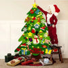 Un enfant déguisé en Père Noël se tient à côté d'un Sapin de Noël en Feutre - 26 Pièces de Décoration + Guirlande Lumineuse - 95cm, décorées avec des feutres.