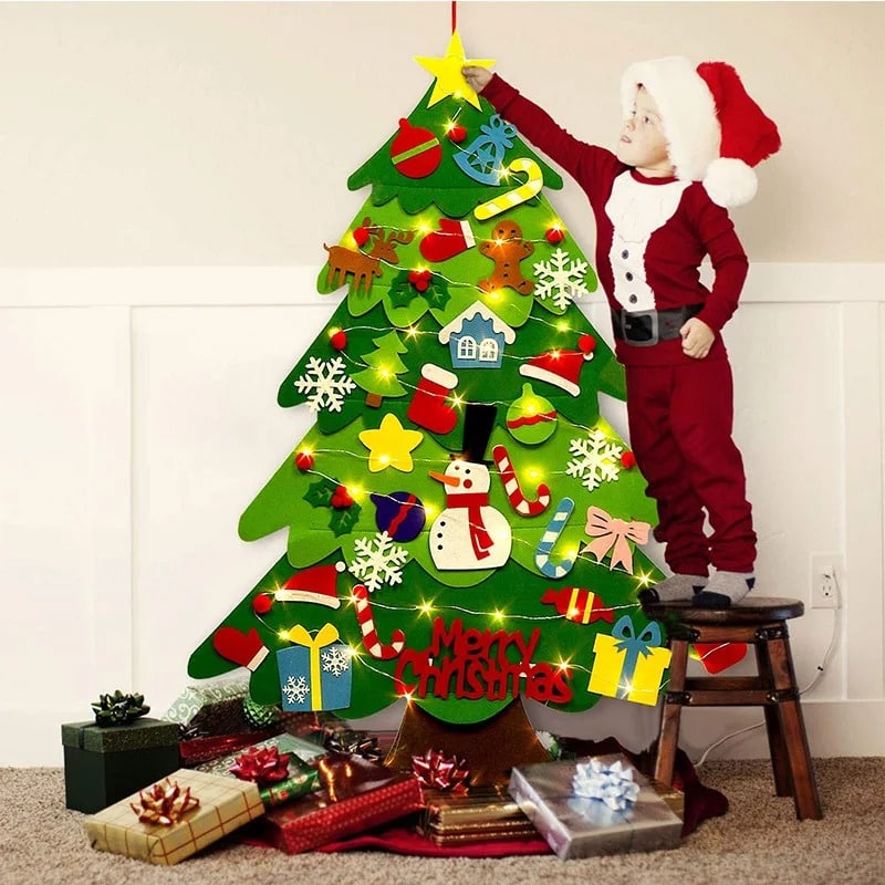 Un enfant déguisé en Père Noël se tient à côté d'un Sapin de Noël en Feutre - 26 Pièces de Décoration + Guirlande Lumineuse - 95cm, décorées avec des feutres.