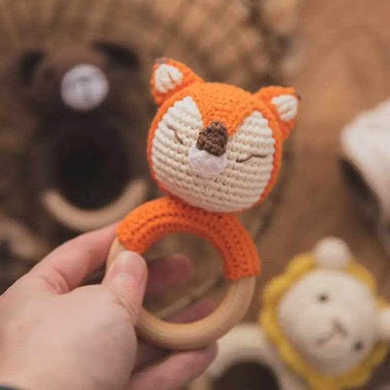 Une main tient un charmant hochet renard Anneaux de dentition Animaux, réalisé au crochet avec un manche en bois, idéal pour les tout-petits. En arrière-plan flou, découvrez le ravissant ensemble composé de hochets ours et lion.