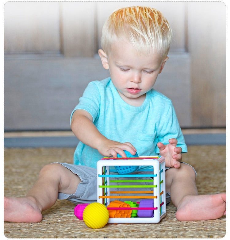 Un tout-petit vêtu d’une chemise bleu clair est assis par terre, explorant avec impatience une Boite à formes avec élastiques – Jeu de motricité colorée.