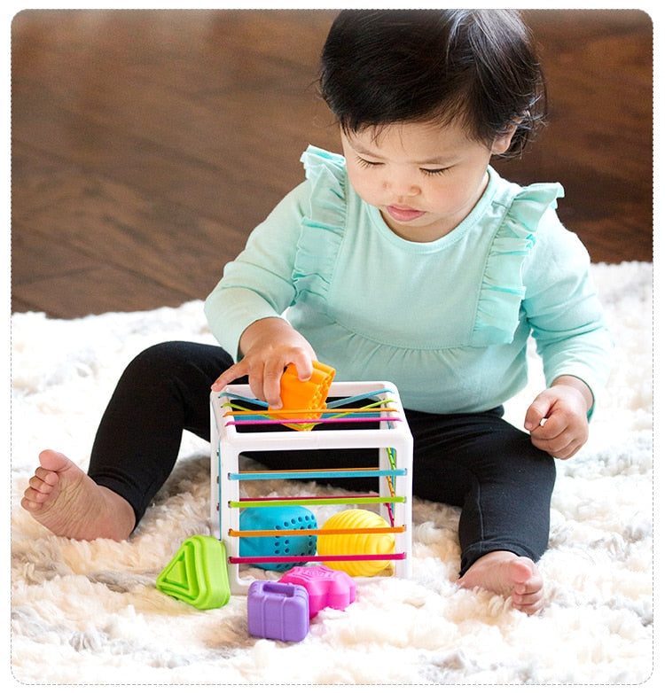 Un tout-petit est assis sur un tapis moelleux et profite de la Boite à formes avec élastiques – Jeu de motricité, un cube jouet décoré de formes géométriques colorées.