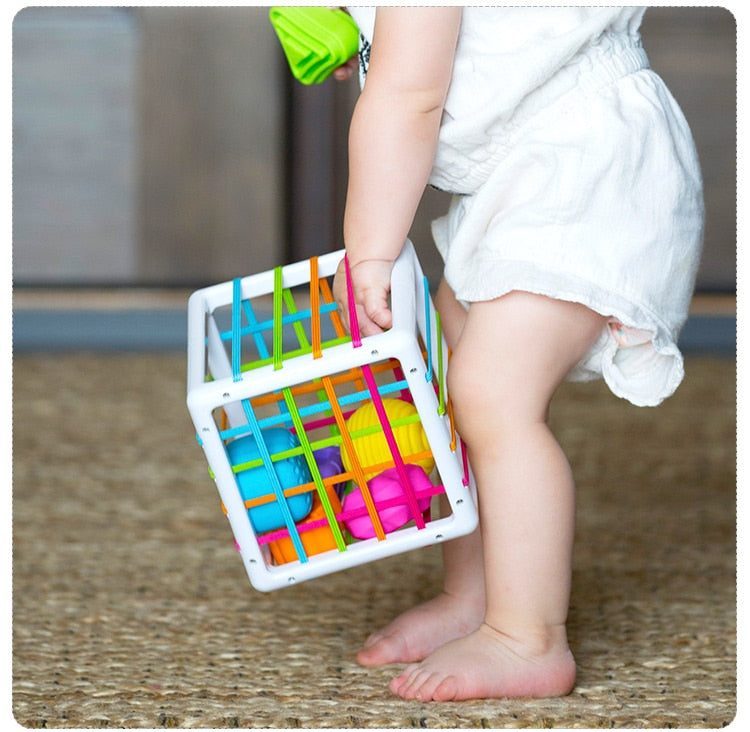 Un bambin vêtu de vêtements blancs se tient pieds nus sur une moquette, tenant une boîte à formes avec élastiques colorée remplie de divers jeux de motricité.