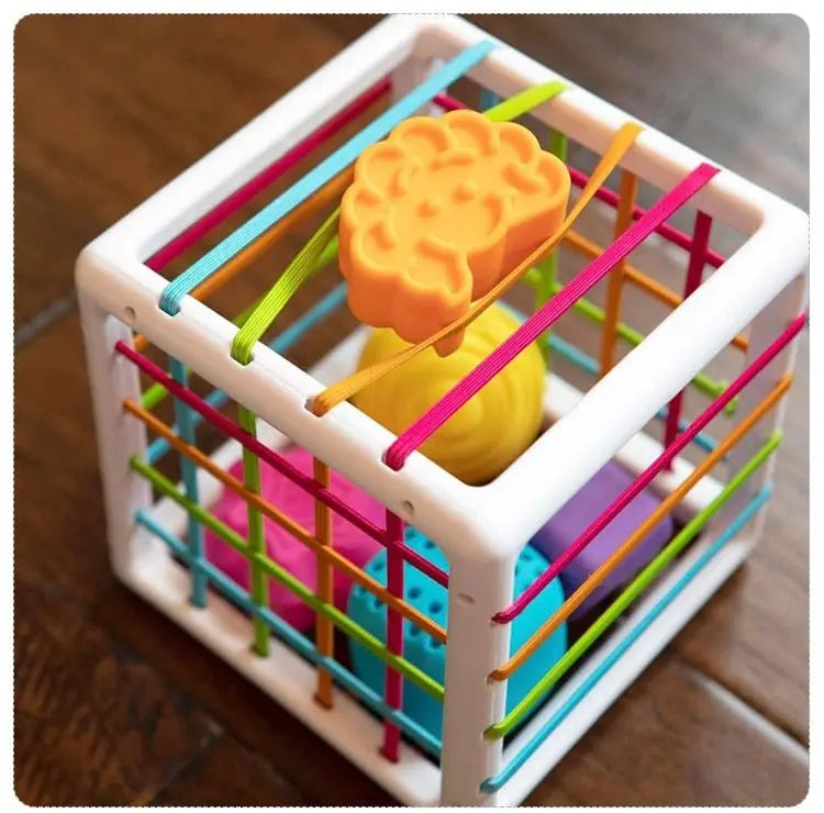 La Boite à formes avec élastiques – Jeu de motricité est un jouet cube vibrant doté d'élastiques et de formes amovibles comme un nuage orange, une sphère jaune et un bloc bleu, idéal pour développer la motricité.