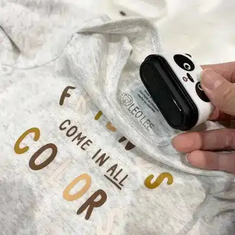 Une main tenant un mètre ruban panda sur une chemise grise sur laquelle est écrit « Les amis viennent de toutes les couleurs » avec des lettres de différentes couleurs, rappelant les dessins ludiques de Mon Tampon Magique Prénom - Pour ne plus perdre d'affaires.